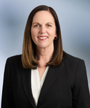 Woman, Anne Seurynck, with black blazer jacket and white blouse smiles at camera in front of blue grey background