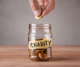 Hand hovers above ready to drop a coin in glass jar full of coins with piece of tape that says charity.