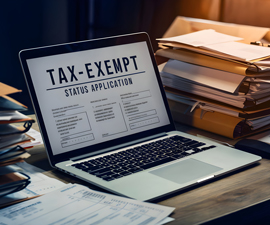 Laptop open on desk surrounded by stacks of files and screen says Tax Exempt Status Application