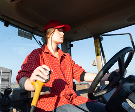 Young teenage woman wearing red flannel shirt and red hat drives inside cab of tractor